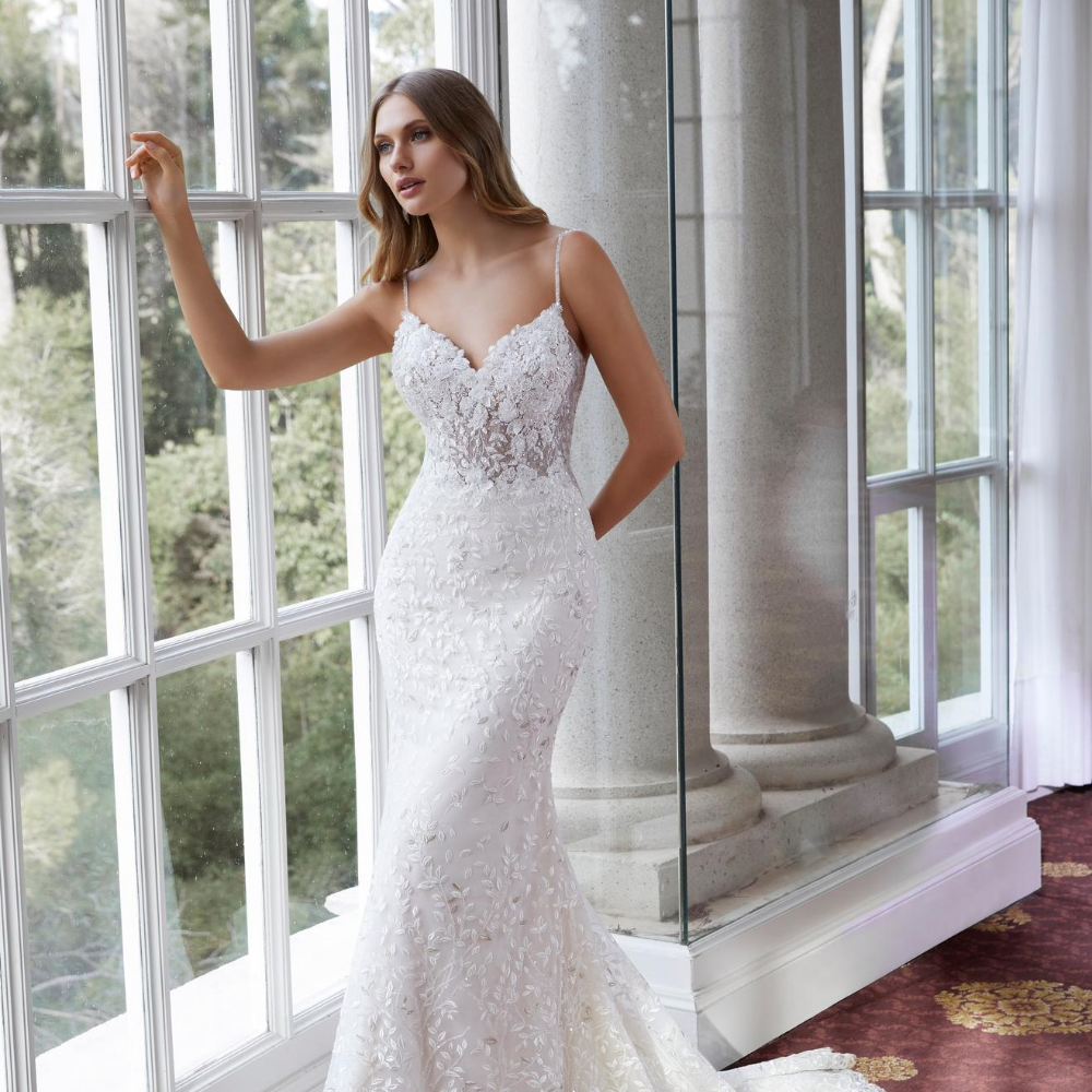 Model stood looking outside floor to ceiling windows in one of our wedding gowns suitable for small weddings, an ivory fit and flare dress with 3D flower appliques and delicate straps